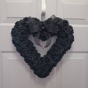 K&ouml;nnte beinhalten: Ein herzf&ouml;rmiger Kranz aus schwarzen Rosen, verziert mit einer schwarzen Schleife aus Organza. Der Kranz h&auml;ngt an einer wei&szlig;en T&uuml;r. Die Rosen sind dicht gepackt, wodurch ein voller, strukturierter Eindruck entsteht. Ein dekoratives Element f&uuml;r die Heimdekoration.