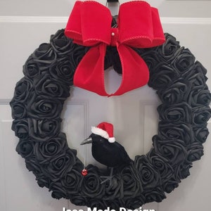 May include: A black wreath made of fabric roses with a red velvet bow and a black crow wearing a red Santa hat. The wreath has a red bell hanging from it. The text "Jess Made Design" is at the bottom of the wreath.