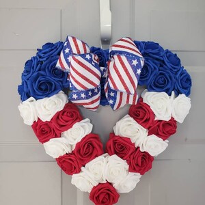 May include: Heart-shaped wreath made of blue, red, and white roses, with a patriotic bow. The wreath is designed with blue roses around the top, red and white roses forming the heart shape, and a bow with red and white stripes and blue stars.