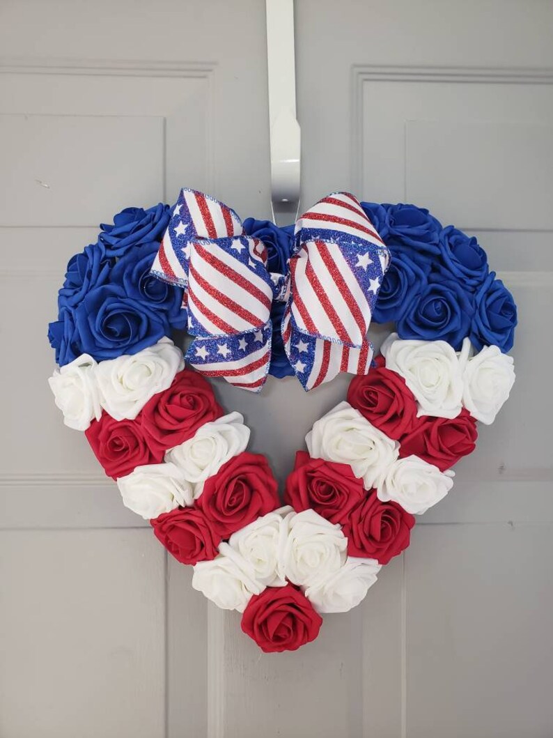 May include: Heart-shaped wreath made of blue, red, and white roses, with a patriotic bow. The wreath is designed with blue roses around the top, red and white roses forming the heart shape, and a bow with red and white stripes and blue stars.
