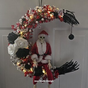 May include: A black and white crescent moon wreath decorated with red and white berries, white and black roses, and a skeleton dressed as Santa Claus. The wreath has white lights and a black ornament hanging from the bottom. The wreath says "Jess Made Design" at the bottom.