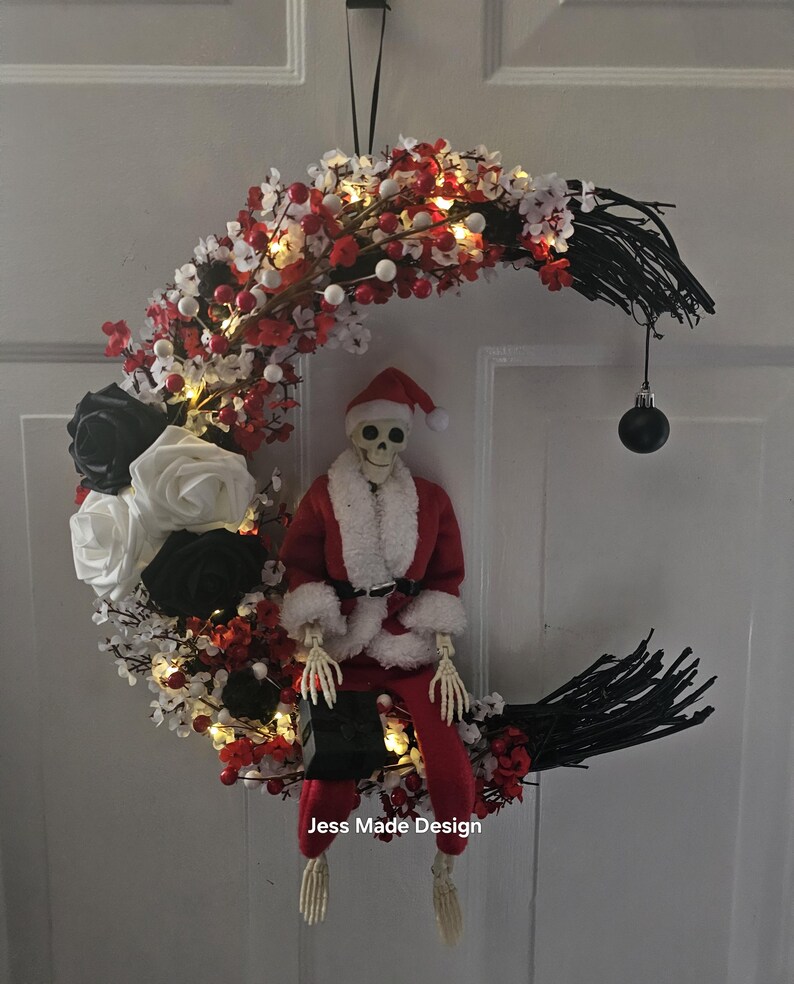 May include: A black and white crescent moon wreath decorated with red and white berries, white and black roses, and a skeleton dressed as Santa Claus. The wreath has white lights and a black ornament hanging from the bottom. The wreath says "Jess Made Design" at the bottom.