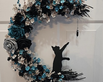 Puede incluir: Una corona en forma de media luna decorada con flores negras, blancas y azul turquesa. Una silueta de gato negro está posada en la luna, y un colgante de cristal cuelga de la corona.