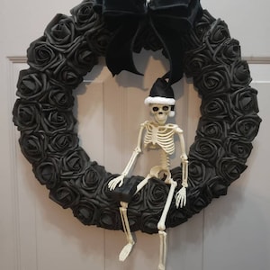 May include: A black wreath made of fabric roses with a black velvet bow and a skeleton wearing a Santa hat sitting on it.