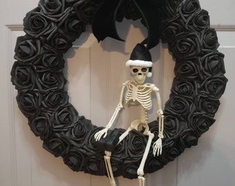 May include: A black wreath made of fabric roses with a black velvet bow and a skeleton wearing a Santa hat sitting on it.