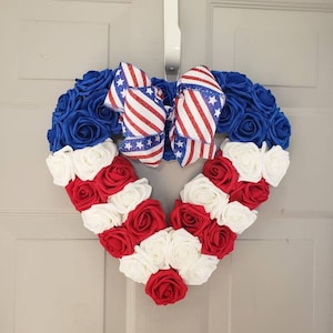 May include: A heart-shaped wreath made of blue, red, and white roses, with a patriotic bow. The wreath is designed with blue roses around the top, red and white roses forming the bottom, and a bow with red and white stripes and blue stars.