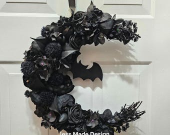 Puede incluir: Una corona de luna negra en forma de media luna decorada con flores negras, hojas y un recorte de murciélago negro. La corona está colgada en una puerta blanca. El texto "Jess Made Design" es visible en la parte inferior de la corona.