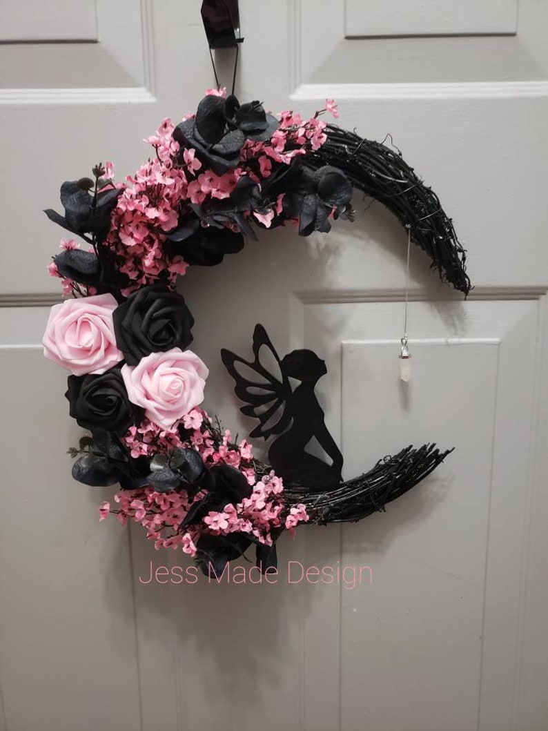 May include: A black and pink crescent moon wreath with a fairy silhouette and pink flowers. The wreath is made of twigs and has a black ribbon hanging from it. The text "Jess Made Design" is visible at the bottom of the image.