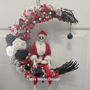 May include: A black and white crescent moon wreath decorated with red and white berries, black and white roses, and a skeleton dressed as Santa Claus holding a gift box. The wreath has a black ribbon for hanging and a black ornament hanging from the bottom. The text "Jess Made Design" is visible at the bottom of the wreath.