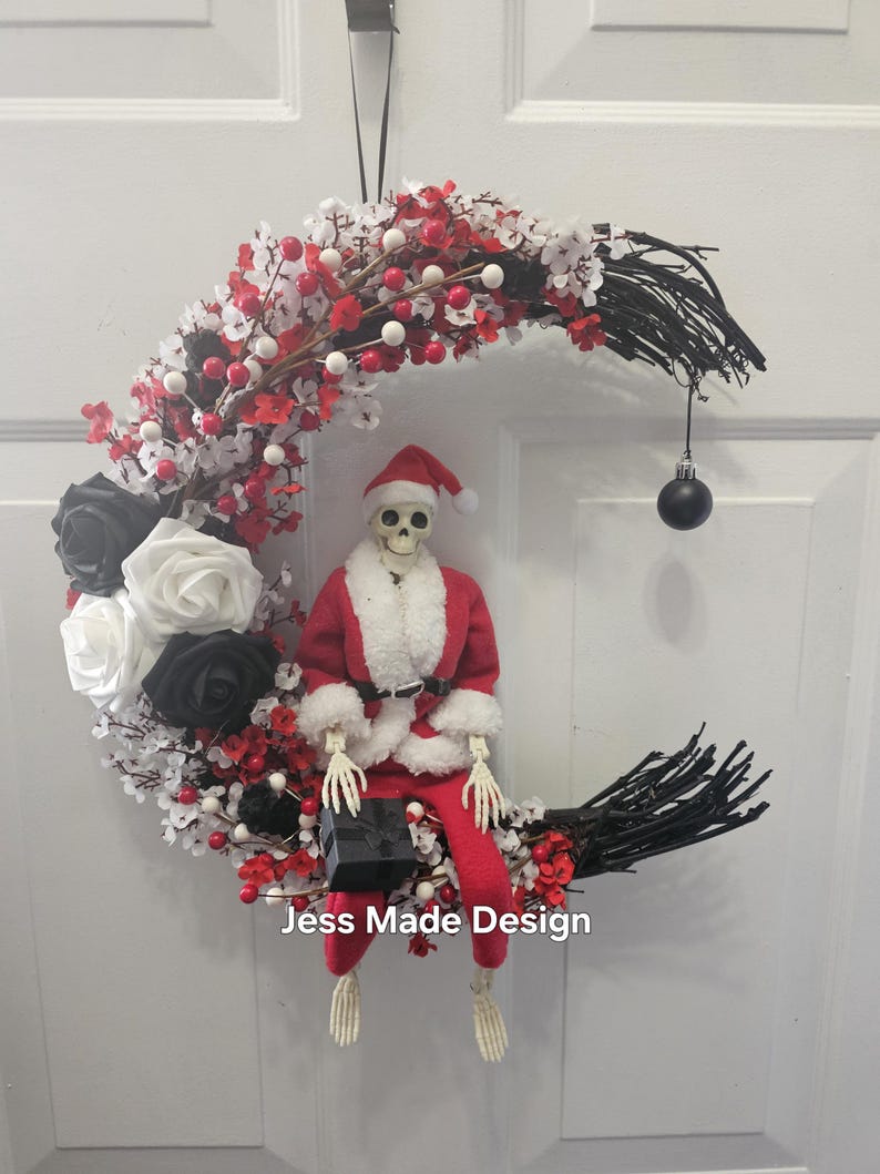 May include: A black and white crescent moon wreath decorated with red and white berries, black and white roses, and a skeleton dressed as Santa Claus holding a gift box. The wreath has a black ribbon for hanging and a black ornament hanging from the bottom. The text "Jess Made Design" is visible at the bottom of the wreath.