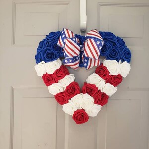 May include: A heart-shaped wreath made of blue, red, and white roses, with a patriotic bow. The wreath is decorated with blue roses at the top, transitioning to red and white roses in the shape of a heart. A bow with red and white stripes and stars is at the top.
