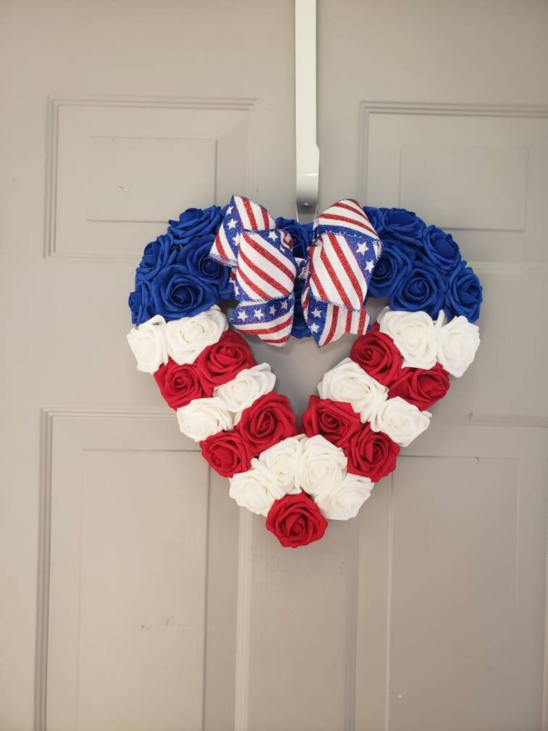 May include: A heart-shaped wreath made of blue, red, and white roses, with a patriotic bow. The wreath is decorated with blue roses at the top, transitioning to red and white roses in the shape of a heart. A bow with red and white stripes and stars is at the top.