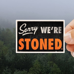 May include: A black and white sign with the text "Sorry We're Stoned" in white and orange lettering. The sign is being held by a hand against a blurry background of green trees.