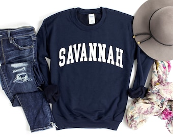 May include: A navy blue crewneck sweatshirt with the word SAVANNAH printed in white block letters across the chest, paired with dark blue distressed denim jeans, a grey wide-brimmed hat, and a floral patterned scarf with pink and green accents.