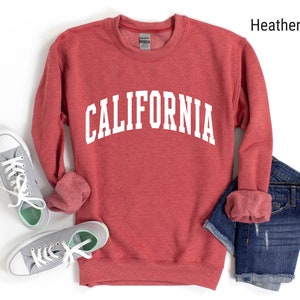May include: A heather red sweatshirt with the word "CALIFORNIA" printed in white on the front. The sweatshirt is shown laid flat on a white background with a pair of grey trainers and blue jeans.