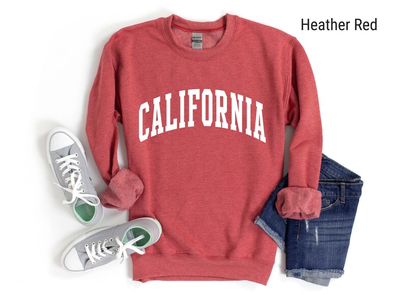 May include: A heather red sweatshirt with the word "CALIFORNIA" printed in white on the front. The sweatshirt is shown laid flat on a white background with a pair of grey trainers and blue jeans.