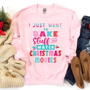 May include: A light pink sweatshirt with the text "I JUST WANT TO BAKE STUFF AND WATCH CHRISTMAS MOVIES" in colorful letters. The sweatshirt is paired with blue jeans, brown flats, and a cream-colored scarf.