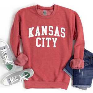 May include: Red sweatshirt with white text that reads "KANSAS CITY". The sweatshirt is shown with a pair of gray sneakers and blue denim shorts.