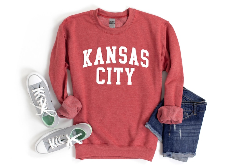 May include: Red sweatshirt with white text that reads "KANSAS CITY". The sweatshirt is shown with a pair of gray sneakers and blue denim shorts.