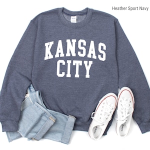 May include: A heather sport navy blue sweatshirt with white text that reads "KANSAS CITY". The sweatshirt is laid flat on a white surface with a pair of white canvas shoes.