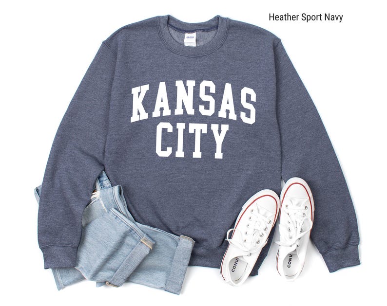 May include: A heather sport navy blue sweatshirt with white text that reads "KANSAS CITY". The sweatshirt is laid flat on a white surface with a pair of white canvas shoes.