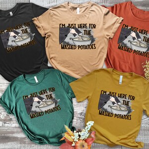 May include: Five different colored t-shirts with a graphic of a hand holding a whisk and mixing mashed potatoes in a bowl. The text on the shirts reads "I'm just here for the mashed potatoes".