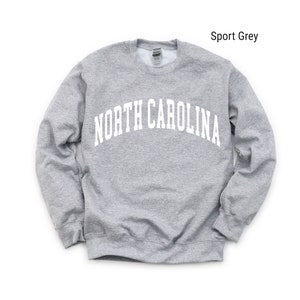 Pode incluir: Uma camisola cinzenta com o texto "NORTH CAROLINA" em branco, em arco sobre o peito. A camisola &eacute; de estilo cl&aacute;ssico de gola redonda.