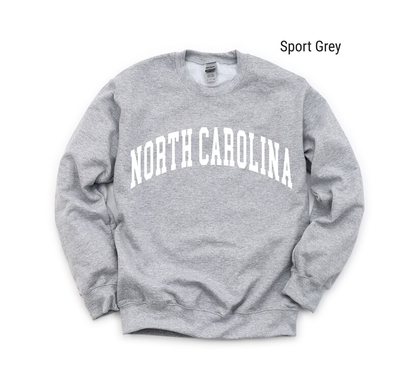 Pode incluir: Uma camisola cinzenta com o texto "NORTH CAROLINA" em branco, em arco sobre o peito. A camisola &eacute; de estilo cl&aacute;ssico de gola redonda.