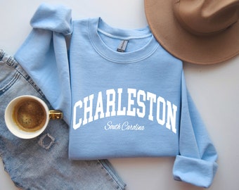 May include: A light blue crewneck sweatshirt featuring the white text CHARLESTON in a bold arched collegiate font, with the words South Carolina written in a smaller cursive script underneath the main city name on the front.