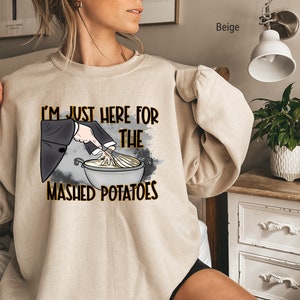 May include: Beige sweatshirt with a graphic of a hand stirring mashed potatoes in a bowl. The text on the sweatshirt reads "I'm just here for the mashed potatoes."
