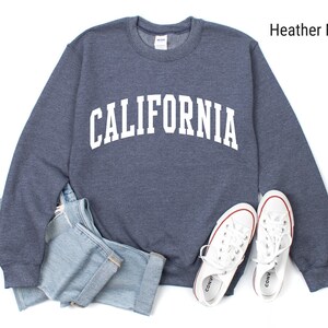 May include: A heather navy blue sweatshirt with the word "CALIFORNIA" printed in white on the front. The sweatshirt is laid flat on a white surface with a pair of white trainers and a pair of blue jeans.