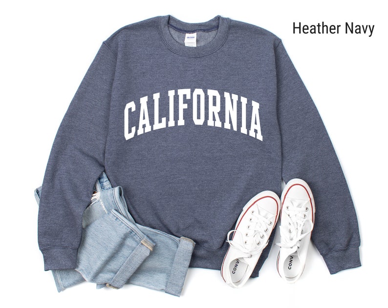 May include: A heather navy blue sweatshirt with the word "CALIFORNIA" printed in white on the front. The sweatshirt is laid flat on a white surface with a pair of white trainers and a pair of blue jeans.