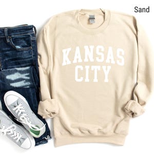 May include: A sand-colored sweatshirt with the text "KANSAS CITY" in white lettering. The sweatshirt is shown with a pair of blue jeans and gray sneakers.