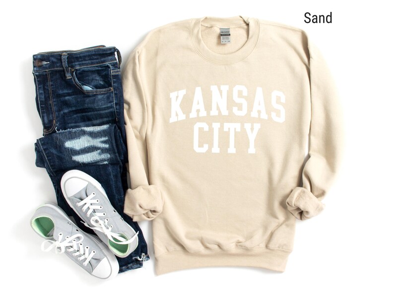 May include: A sand-colored sweatshirt with the text "KANSAS CITY" in white lettering. The sweatshirt is shown with a pair of blue jeans and gray sneakers.