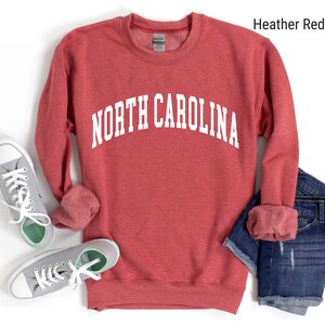 Pode incluir: Camisola de cor vermelha com o texto "NORTH CAROLINA" em branco. A camisola est&aacute; dobrada e colocada sobre uma superf&iacute;cie branca com um par de t&eacute;nis cinzentos e cal&ccedil;&otilde;es de ganga azuis.