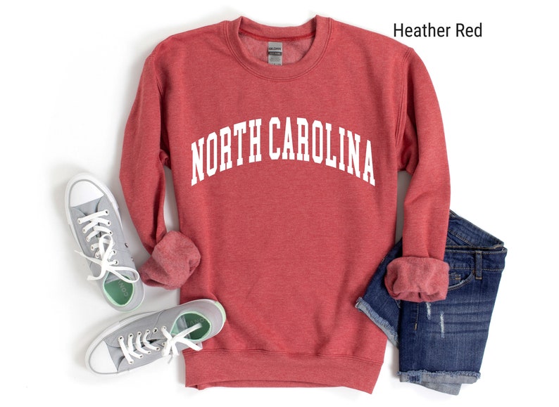 Pode incluir: Camisola de cor vermelha com o texto "NORTH CAROLINA" em branco. A camisola est&aacute; dobrada e colocada sobre uma superf&iacute;cie branca com um par de t&eacute;nis cinzentos e cal&ccedil;&otilde;es de ganga azuis.