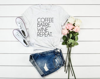 May include: A gray t-shirt with the text "COFFEE BARRE. WINE. REPEAT." on it. The shirt is paired with denim shorts and white sneakers. A bouquet of pink and white roses is in the background.