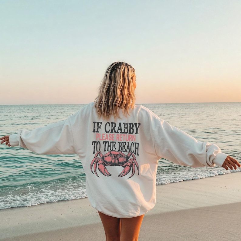 May include: A white sweatshirt with the text "IF CRABBY PLEASE RETURN TO THE BEACH" and a pink crab graphic. The sweatshirt is worn on a beach with the ocean in the background.