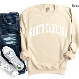 Pode incluir: Camisola de moletom bege com o texto "NORTH CAROLINA" em letras brancas. A camisola est&aacute; colocada sobre um fundo branco com um par de cal&ccedil;as de ganga azuis e t&eacute;nis cinzentos.