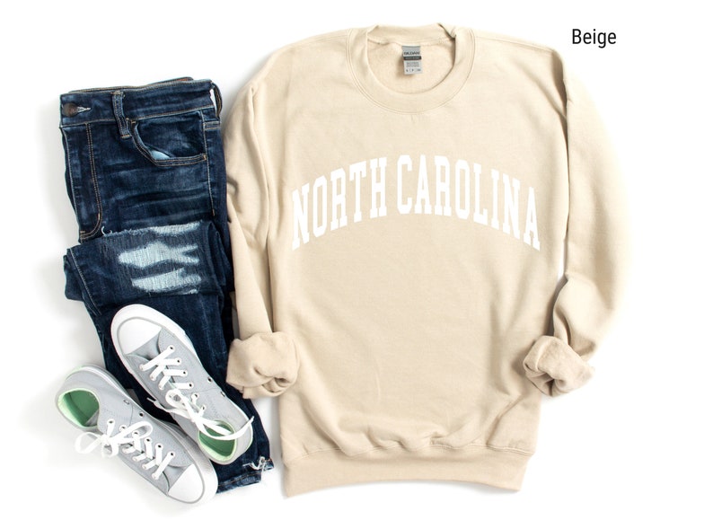 Pode incluir: Camisola de moletom bege com o texto "NORTH CAROLINA" em letras brancas. A camisola est&aacute; colocada sobre um fundo branco com um par de cal&ccedil;as de ganga azuis e t&eacute;nis cinzentos.