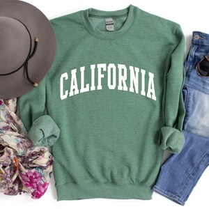 May include: A green sweatshirt with the word "CALIFORNIA" printed in white on the front. The sweatshirt is laid flat on a white background with a hat, scarf, and jeans.