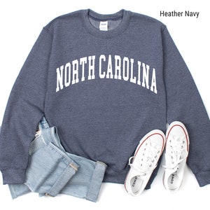 Pode incluir: Uma camisola azul-marinho desbotada com o texto "NORTH CAROLINA" em branco, em letras arqueadas. A camisola est&aacute; colocada sobre um fundo branco com um par de t&eacute;nis brancos e cal&ccedil;as de ganga azuis.