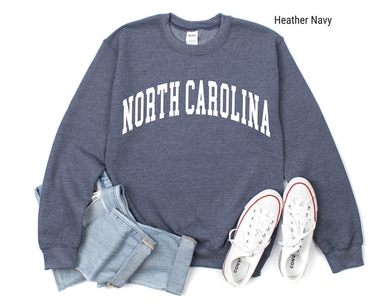 Pode incluir: Uma camisola azul-marinho desbotada com o texto "NORTH CAROLINA" em branco, em letras arqueadas. A camisola est&aacute; colocada sobre um fundo branco com um par de t&eacute;nis brancos e cal&ccedil;as de ganga azuis.