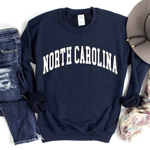 Pode incluir: Uma camisola azul-marinho com o texto "NORTH CAROLINA" em branco, combinada com cal&ccedil;as de ganga azuis desgastadas, um len&ccedil;o floral e um chap&eacute;u fedora cinzento.