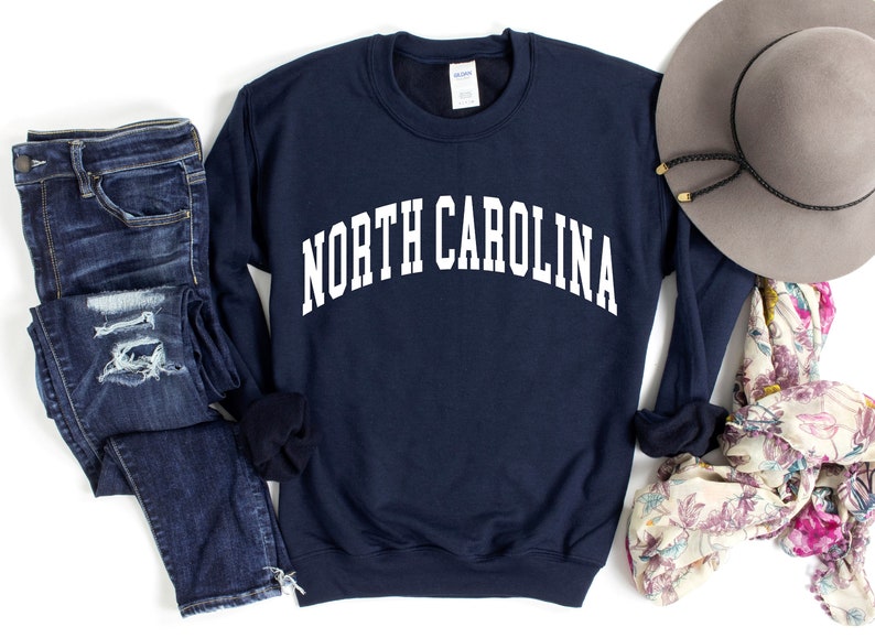 Pode incluir: Uma camisola azul-marinho com o texto "NORTH CAROLINA" em branco, combinada com cal&ccedil;as de ganga azuis desgastadas, um len&ccedil;o floral e um chap&eacute;u fedora cinzento.