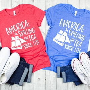 May include: Two t-shirts with a red and blue color scheme. The shirts feature a white graphic of a ship with the text "America: Spilling the Tea Since 1773".