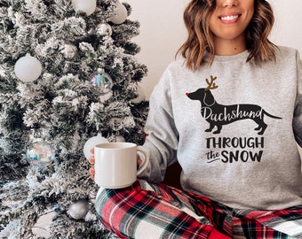 May include: A gray sweatshirt with a black dachshund wearing a red nose and reindeer antlers. The text "Dachshund Through the Snow" is printed below the dog.
