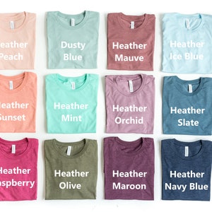 May include: Twelve folded t-shirts in various heather colors, including peach, dusty blue, mauve, ice blue, sunset, mint, orchid, slate, raspberry, olive, maroon, and navy blue.