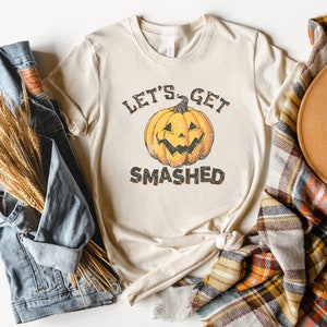 May include: A beige t-shirt with a vintage-style graphic of a pumpkin with the text "Let's Get Smashed". The shirt is displayed on a white background with a denim jacket, a plaid blanket, and a brown hat.