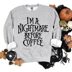 Peut inclure: Un sweat-shirt gris avec le texte "I'm a Nightmare Before Coffee" imprim&eacute; en noir. Le sweat-shirt est photographi&eacute; sur un fond blanc avec des d&eacute;corations d'Halloween, notamment des citrouilles, des toiles d'araign&eacute;es et des feuilles.